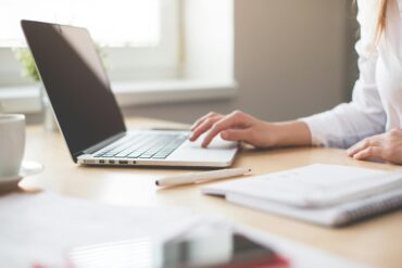 A businesswoman typing on a laptop at a bright indoor workspace with notebooks and a pen.